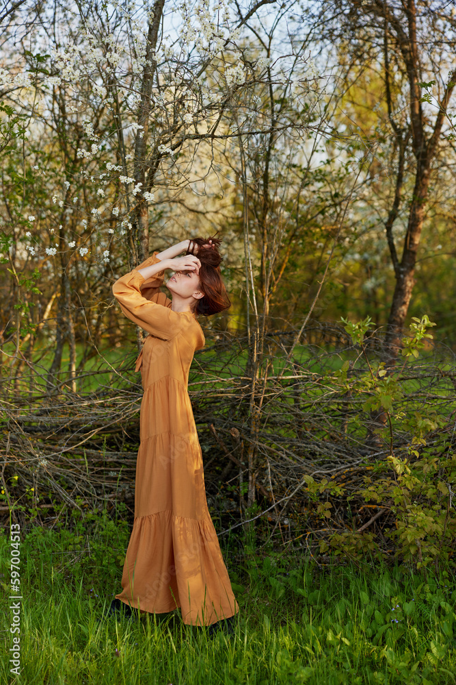an elegant, sophisticated woman poses standing near a wicker fence in a dacha in a long orange dress, straightening her hair with her hands an elegant, sophisticated woman poses standing near a wicker fence in a dacha in a long orange dress, straightening her hair with her hands