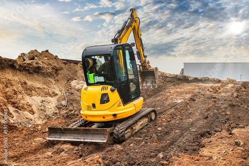 Mini excavator at the construction site on the edge of a pit against a cloudy blue sky. Compact construction equipment for earthworks. An indispensable assistant for earthworks.