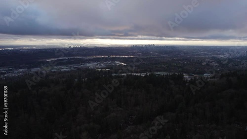 Aerial cityscape view of Vancouver, BC during a cloudy sunset with a mountain forest in the foreground