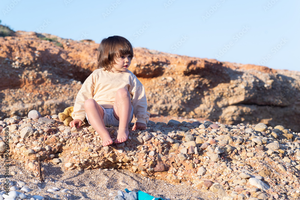 Two years old girl sitting and  relaxing on a rock formation with barefoot on a pebble beach at sunset  in summer. 