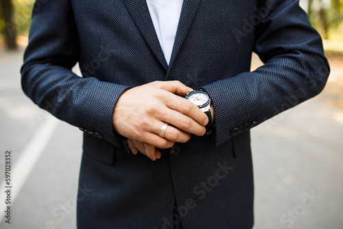 successful married businessman in a dark gray suit pays attention to the watch on his hand. The man pays attention to the time while standing on the road. Close-up