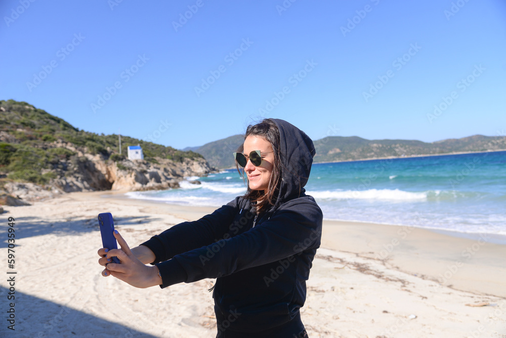Cheerful young woman taking a selfie while standing next to the sea. Young girl smiling happily looking at her mobile phone. Outdoors summer sun fun.