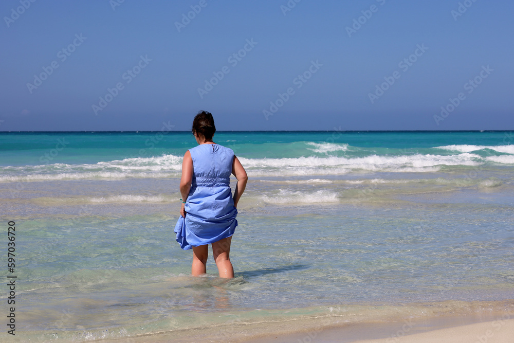 Woman in blue dress going to azure sea water. Travel and beach vacation on sandy coast