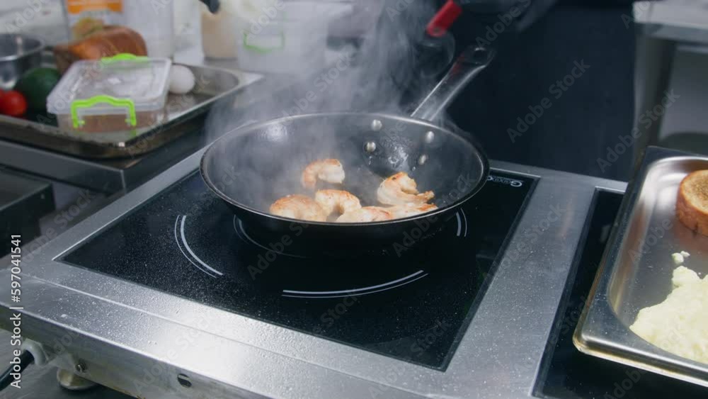 Male cook fries and stirs shrimp in frying pan, pours cooking oil. The chef cooks delicious seafood dish. Cooking process on restaurant or cafe kitchen. Professional gourmet cuisine. Public catering.