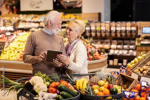 A senior couple is following list on tablet and purchasing groceries at the supermarket.