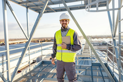A heavy industry worker standing on silo with tablet in his hands and smiling at the camera.