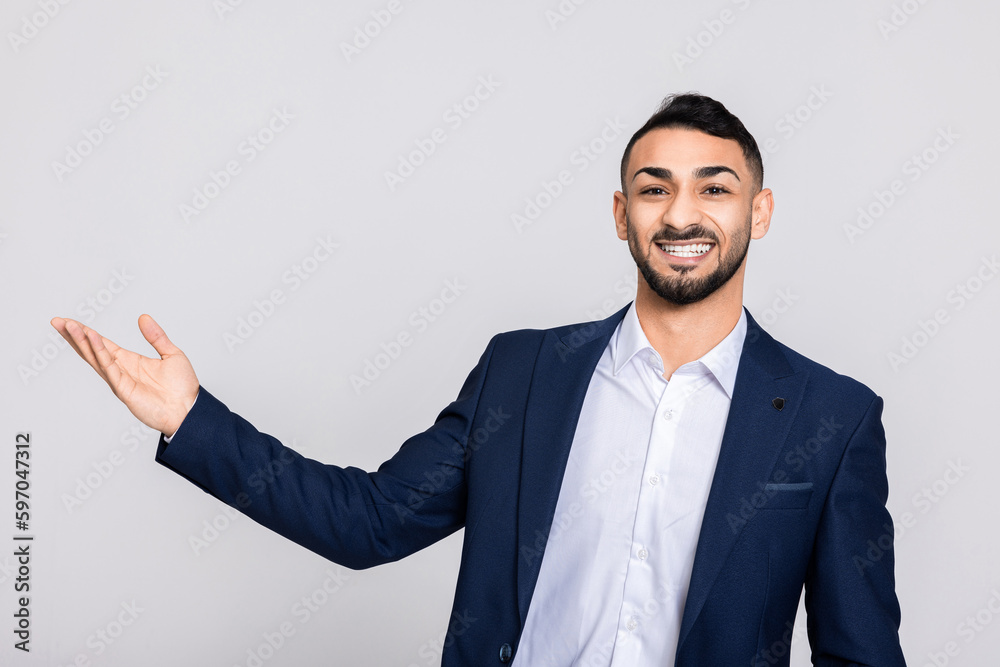 Middle eastern young attractive man buisnessman with beard looking at camera smiling pointing at something talking at camera standing over grey background. Middle eastern young attractive man buisnessman with beard looking at camera smiling pointing at something talking at camera standing over grey background.