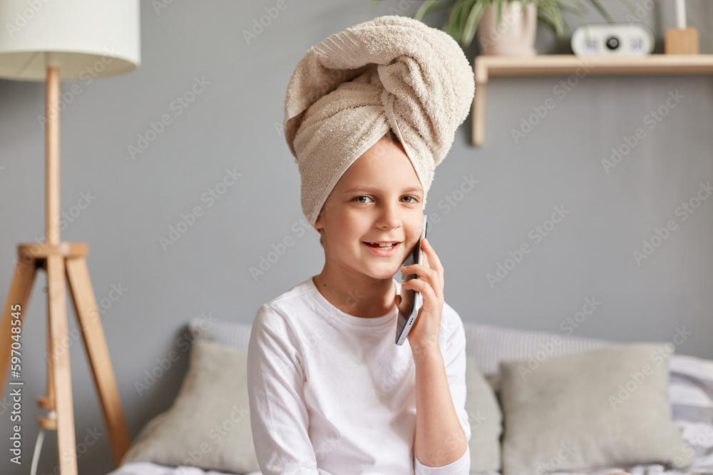 Attractive kid girl talking on phone sitting on bed after bathroom, enjoy, relax, listen to talk of friend, wearing white shirt and towel posing at home. Attractive kid girl talking on phone sitting on bed after bathroom, enjoy, relax, listen to talk of friend, wearing white shirt and towel posing at home.