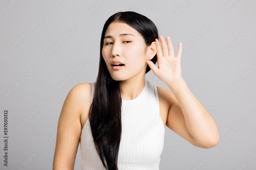 Young curious inquisitive asian woman girl lady standing on grey background in studio isolated holding hand near ears trying to overhear new information gossip about somebody or discounts. Young curious inquisitive asian woman girl lady standing on grey background in studio isolated holding hand near ears trying to overhear new information gossip about somebody or discounts.