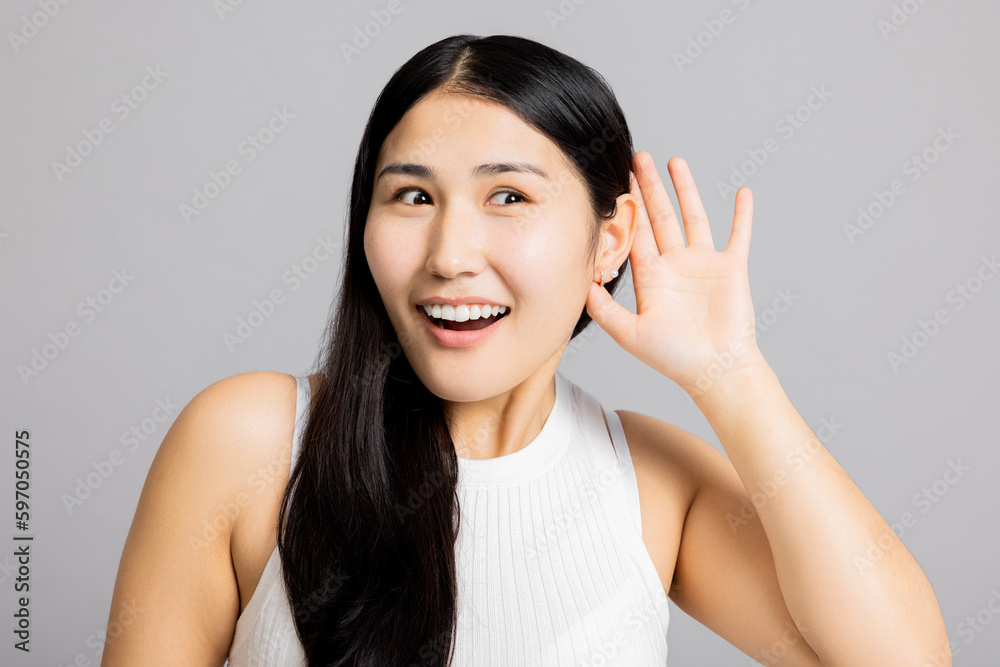 Curious surprised asian girl lady woman holds hand near ear listens new interesting gossip news tries to overhear stands on grey isolated background in studio wearing basic white t-shirt. Curious surprised asian girl lady woman holds hand near ear listens new interesting gossip news tries to overhear stands on grey isolated background in studio wearing basic white t-shirt.