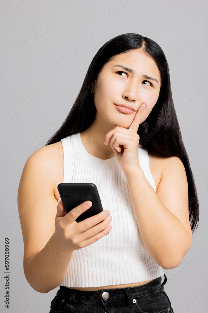Portrait of young cute successful cheerful asian korean woman thinkingshowing thoughtful expressions emotions girl in bad mood isolated on grey background holding cellphone showing emotions at camera. Portrait of young cute successful cheerful asian korean woman thinkingshowing thoughtful expressions emotions girl in bad mood isolated on grey background holding cellphone showing emotions at camera.