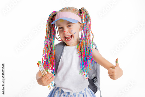 Delightful small pupil student with colorful braids shows thumb up holds painting brushes in hand wears tennis cap and grey backpack ready to go to school pretty girl in good mood.