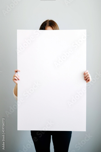 Woman with big paper sheet in her hands, studio shot. Mockup with copy space, no face. Girl covers her face with a white sheet of paper, presenting something, space for text. AI generated 