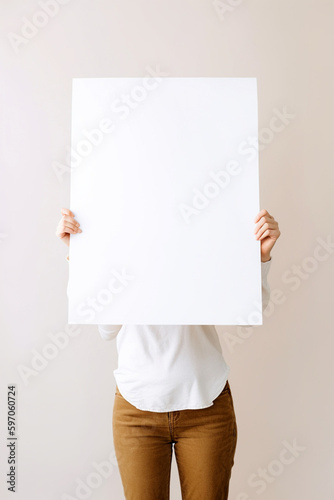 Woman with big paper sheet in her hands, studio shot. Mockup with copy space, no face. Girl covers her face with a white sheet of paper, presenting something, space for text. AI generated image