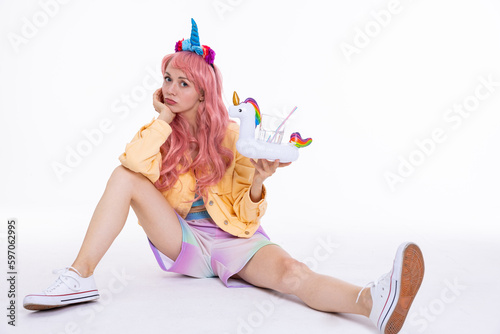 Stylish young girl in pink wig and unicorn blue headband holding water in glas in inflatable white unicorn toy sitting on floor posing for camera in bad mood boring.