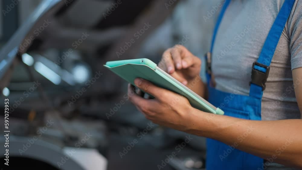 Closeup digital tablet in male hands at background of open car hood in garage. Unrecognizable Caucasian auto mechanic checking vehicle with application indoors at service station Closeup digital tablet in male hands at background of open car hood in garage. Unrecognizable Caucasian auto mechanic checking vehicle with application indoors at service station