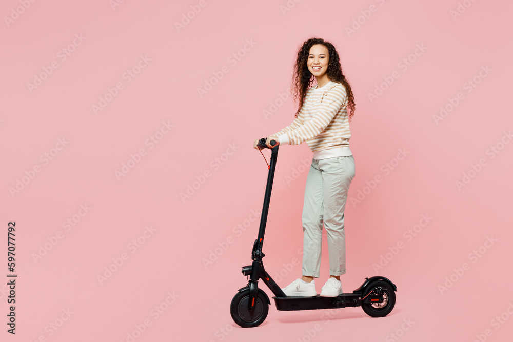 Full body side view smiling happy fun young woman of African American ethnicity wear light casual clothes ridint electrc scooter look camera isolated on plain pastel pink background studio portrait. Full body side view smiling happy fun young woman of African American ethnicity wear light casual clothes ridint electrc scooter look camera isolated on plain pastel pink background studio portrait.