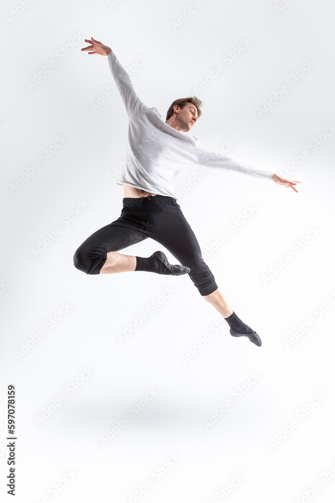 One Active Professional Caucasian Handsome Young Athlete Man Posing in Flying Ballet Pose with Lifted Hands in White Shirt On White.