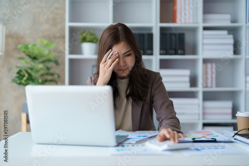 Portrait of a young Asian woman showing a serious face as she uses her calculator, financial documents and computer laptop on her desk in the early morning hours. © Jirapong