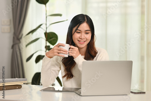 Attractive young asian woman drinking coffee and watching video clip or reading email on laptop. People, technology and lifestyle