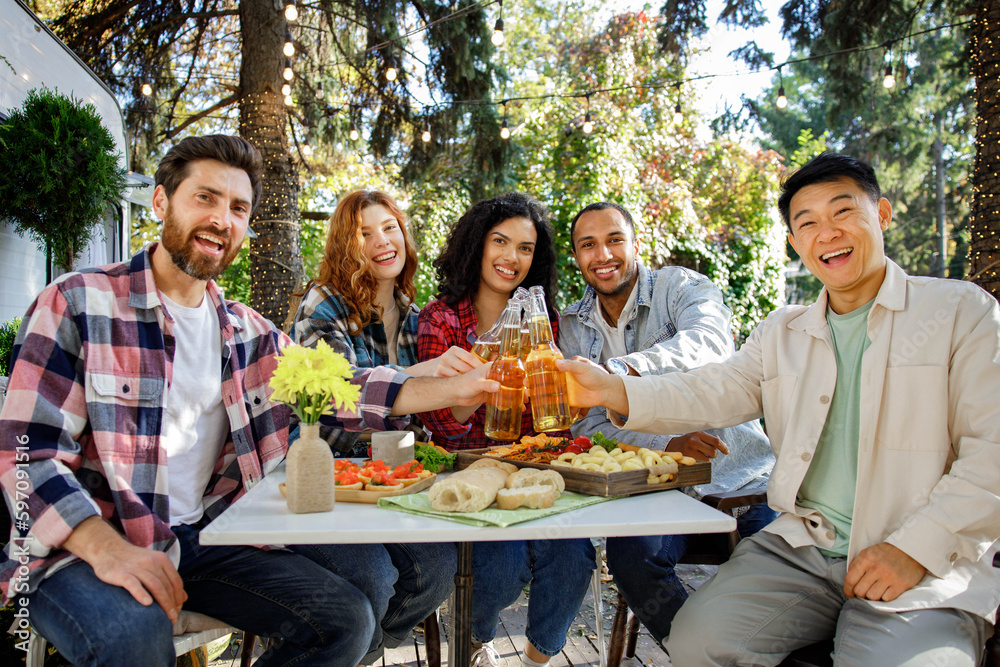 A cheerful multinational company is having fun on a picnic. Multisex friends sitting at the table outdoors celebrating eating and drinking drinks. Look at the camera. A cheerful multinational company is having fun on a picnic. Multisex friends sitting at the table outdoors celebrating eating and drinking drinks. Look at the camera.