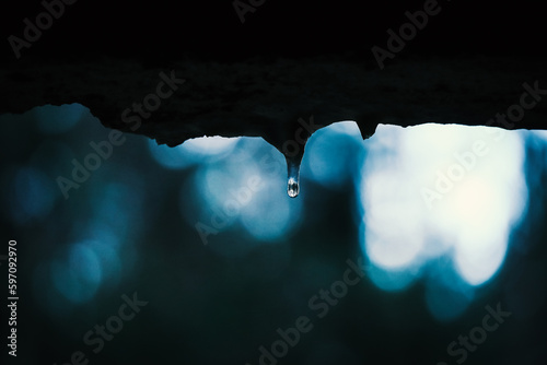 Small water drop with reflection of a forest on icicle formed on the edge of the wall window with visible blurred forest on the background in evening twilight