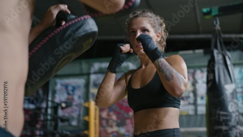 Female muay thai fighter training with coach in gym wearing black boxing bandages and athletic clothing. Caucasian woman kickboxer working out punches and kicks with partner