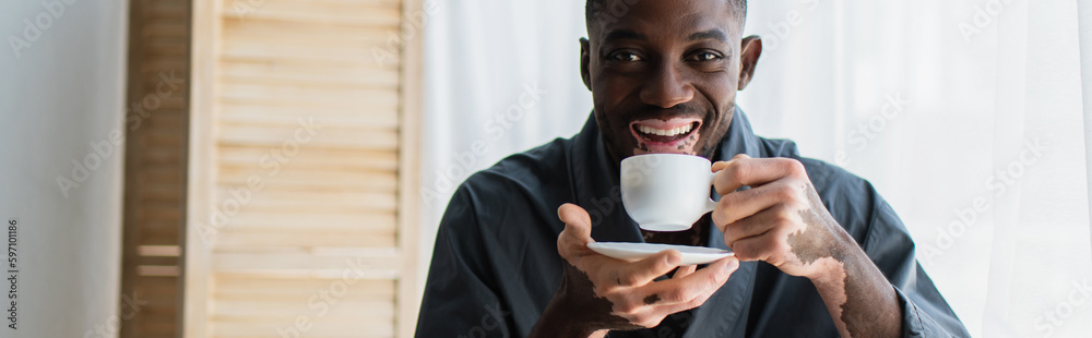 Positive african american man with vitiligo holding coffee cup and looking at camera at home, banner. Positive african american man with vitiligo holding coffee cup and looking at camera at home, banner.