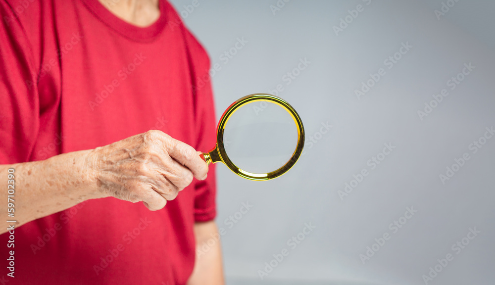 Senior woman wearing a red casual holding a magnifying glass while standing against a gray background Senior woman wearing a red casual holding a magnifying glass while standing against a gray background
