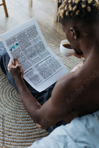 Shirtless african american man with vitiligo reading newspaper and drinking coffee at home.