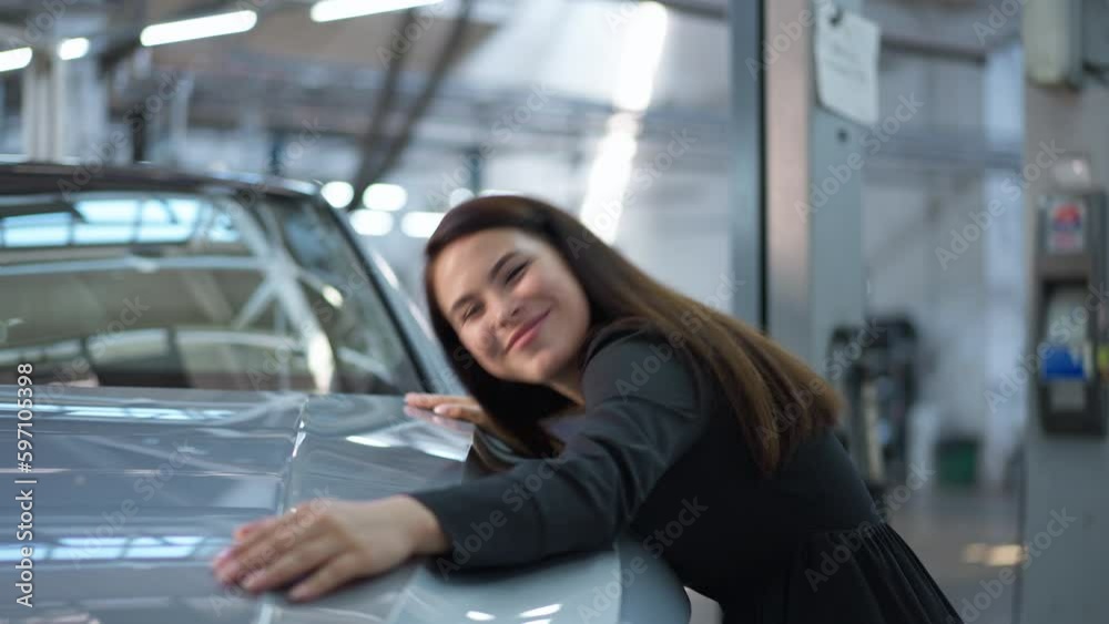 Satisfied young lady hugging car hood smiling and looking at camera in slow motion. Side view portrait of happy new automobile owner posing in dealership indoors Satisfied young lady hugging car hood smiling and looking at camera in slow motion. Side view portrait of happy new automobile owner posing in dealership indoors