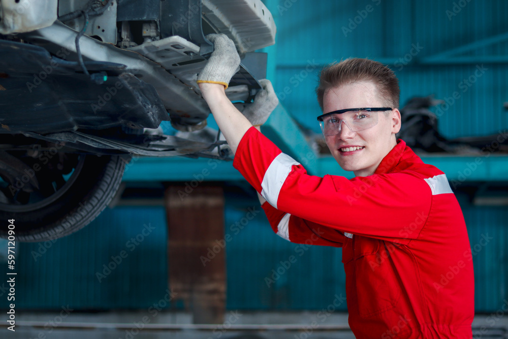 Smiling handsome mechanic man in red uniform work with lifted vehicle, auto mechanic checking wheel at garage, technician check, repair and maintenance customer car automobile at repair service shop.