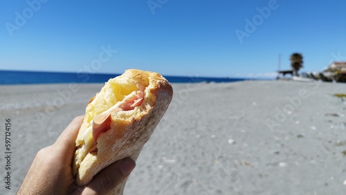 Person eats a sandwich at the beach looking at the blue sea under a beautiful blue sky