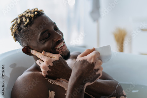 Overjoyed african american man with vitiligo using wireless earphone and holding coffee cup in bathtub at home.