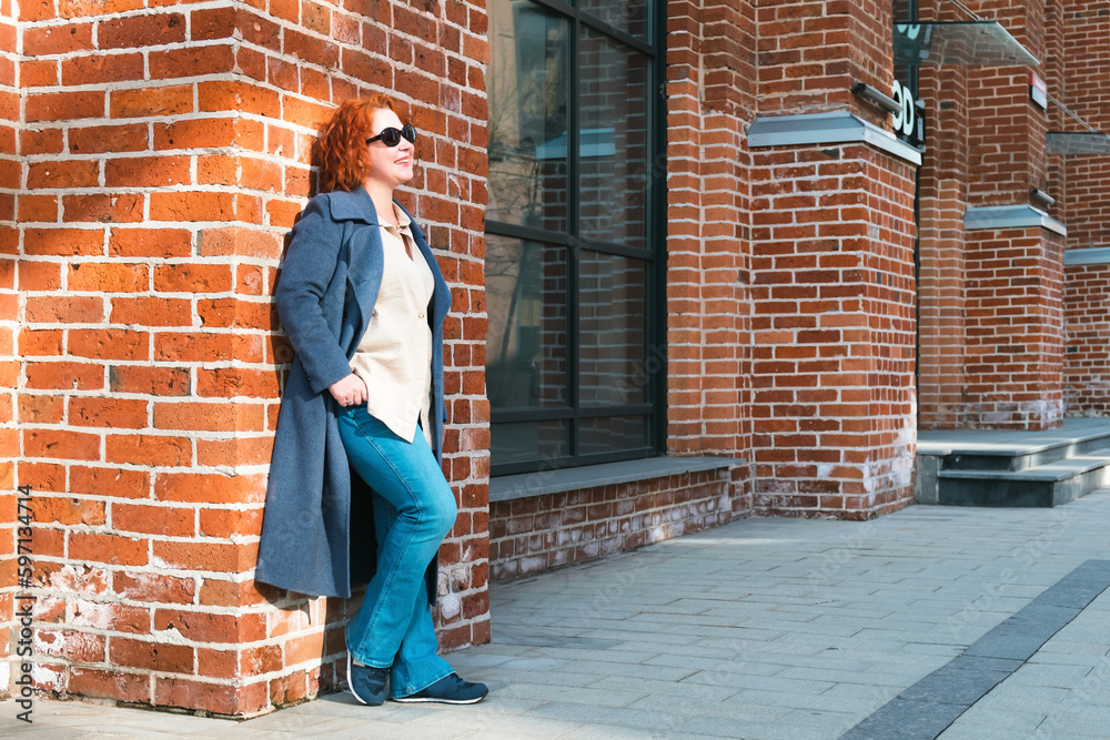 Attractive red-haired woman in a blue coat, jeans stands outside a brick building. Woman in sunglasses looks up and smiles. Portrait of a woman in full growth Attractive red-haired woman in a blue coat, jeans stands outside a brick building. Woman in sunglasses looks up and smiles. Portrait of a woman in full growth