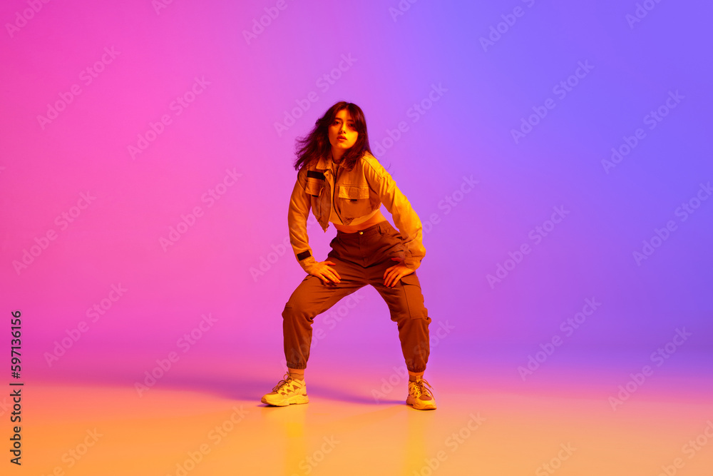 Active, artistic young woman, hip hop dancer in sport style clothes dancing against gradient pink purple background in neon light. Concept of contemporary dance, youth, hobby, action and motion