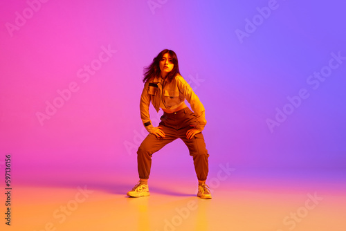 Active, artistic young woman, hip hop dancer in sport style clothes dancing against gradient pink purple background in neon light. Concept of contemporary dance, youth, hobby, action and motion