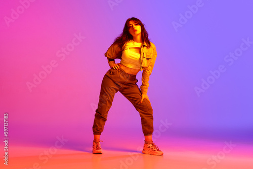Artistic, beautiful, young girl dancing hip-hop against gradient pink purple background in neon light. Model in casual clothes. Concept of contemporary dance, youth, hobby, action and motion