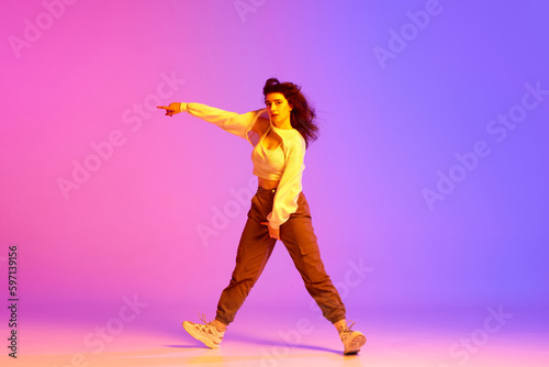 Dynamic image of young sportive girl, female hip-hop dancer training against gradient pink purple background in neon light. Concept of contemporary dance, youth, hobby, action and motion