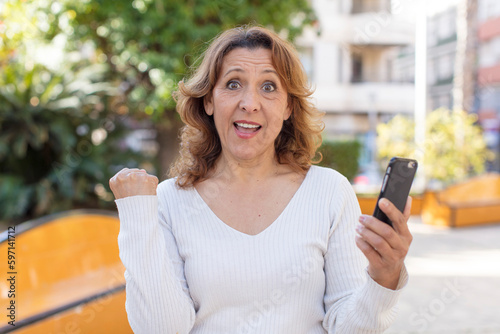 middle age pretty woman feeling shocked,laughing and celebrating success. using her smartphone