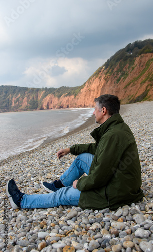A man sits on a beach in front of a cliff, staring out to sea. Concept image, he ponders his retirement, finances, health and pensions. He is isolated and alone. portrait view, copy space. Editorial.