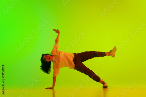 Artistic, flexible, talented young girl, hip-hop dancer performing against gradient green yellow background in neon light. Concept of contemporary dance, youth, hobby, action and motion