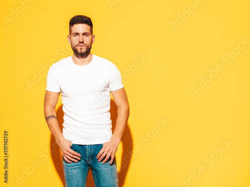 Portrait of handsome smiling stylish hipster lambersexual model. Sexy man dressed in white T-shirt and jeans. Fashion male isolated on yellow wall in studio