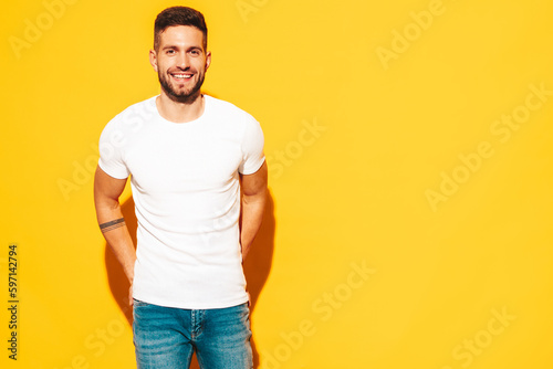 Portrait of handsome smiling stylish hipster lambersexual model. Sexy man dressed in white T-shirt and jeans. Fashion male isolated on yellow wall in studio