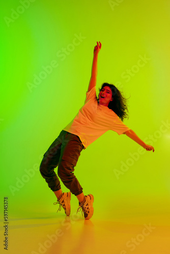 Fototapeta Naklejka Na Ścianę i Meble -  Stylish young girl in sport style clothes dancing hip-hop, training against gradient green yellow background in neon light. Concept of contemporary dance, youth, hobby, action and motion