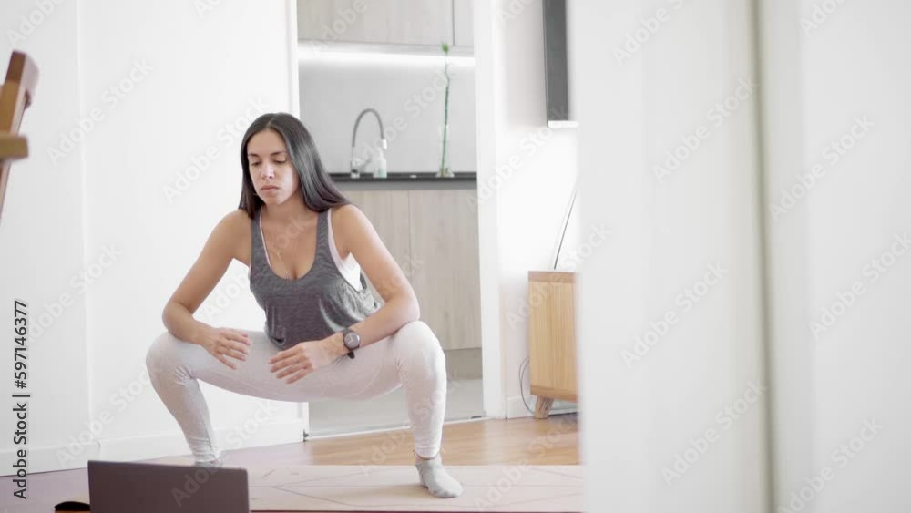 Tilt down and tilt up of a young latin woman doing a sumo squat in her living room while following an online class from her laptop. Copy space. Tilt down and tilt up of a young latin woman doing a sumo squat in her living room while following an online class from her laptop. Copy space.