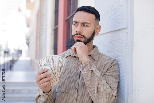 young  adult man smiling with a happy, confident expression with hand on chin. dollar banknotes concept