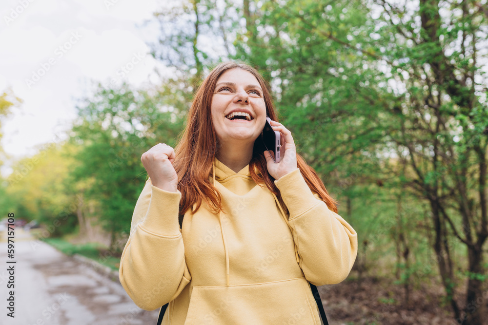 Portrait of a satisfied young woman holding mobile phone and celebrating outdoors. Smiling beautiful women talking on smartphone on nature background Portrait of a satisfied young woman holding mobile phone and celebrating outdoors. Smiling beautiful women talking on smartphone on nature background