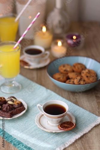 Plate of chocolate pralines, bowl of cookies, cups of tea, glasses of juice and lit candles on the table. Selective focus.