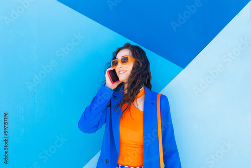 Happy fashionable woman wearing sunglasses talking on smart phone in front of blue wall